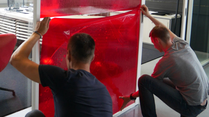Kaksi teippaajaa asentavat läpinäkyviä värikalvoja toimiston lasiseiniin näkösuojaksi